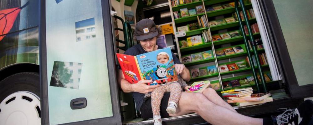 En pappa sitter i dörröppningen i en bokbuss och läser högt för sitt barn. 