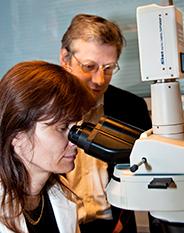 En kvinna som tittar i ett mikroskop. En man står vid sidan om. 