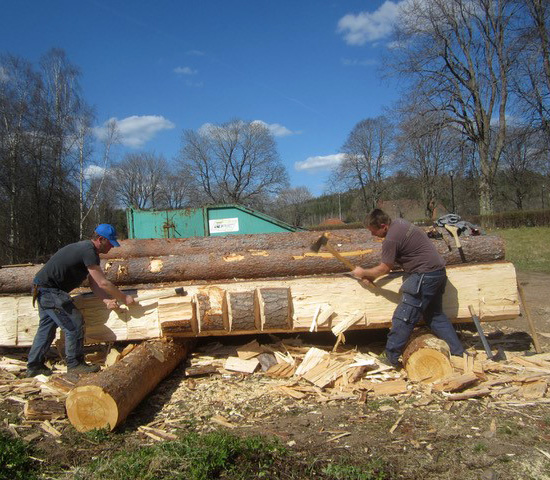 Två personer yxhugger en tjock, liggande stock vid restaureringen.