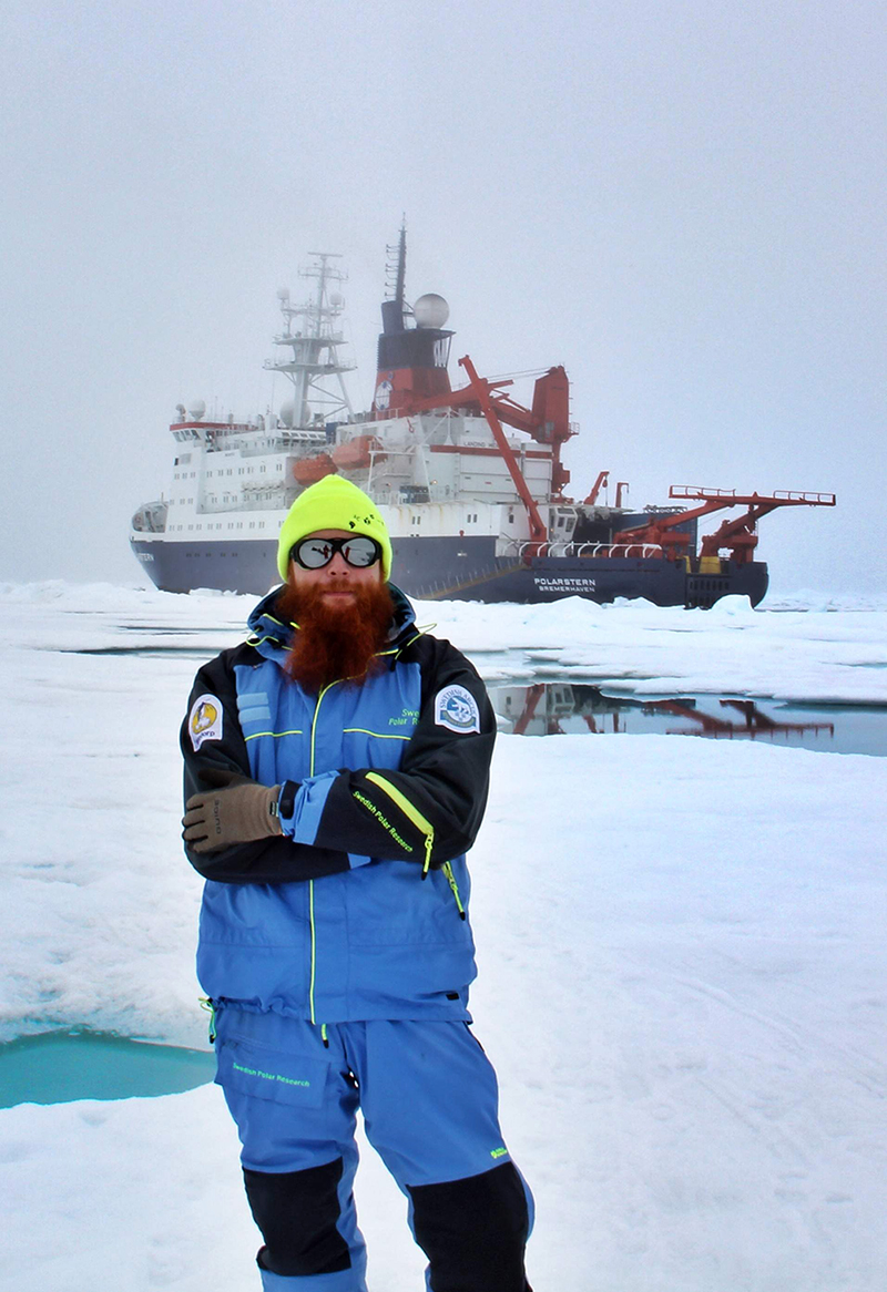 En väl påpälsad Adam Ulfsbo i polarlandskap med isbrytare i bakgrunden.