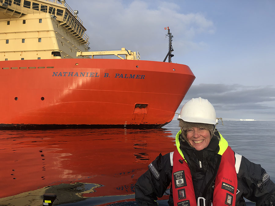 Portätt av leende Anna Wåhlin med hjälm, sitter i båt, isbrytare i bakgrund.