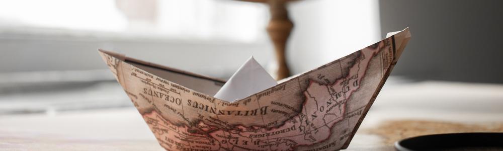 A paper boat made by folding a map is placed on a table in front of a globe.