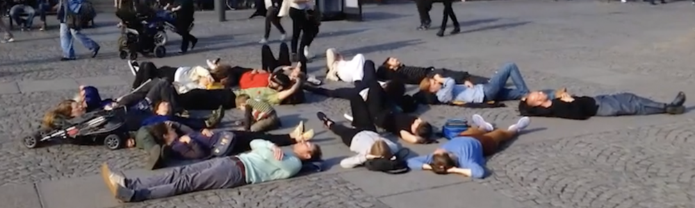 Performance där människor ligger på marken på ett torg. I bakgrunden passerar andra personer och det finns utomhusserveringar.
