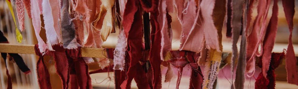 Tygremsor i olika nyanser av rött och rosa hänger över en vävstol.