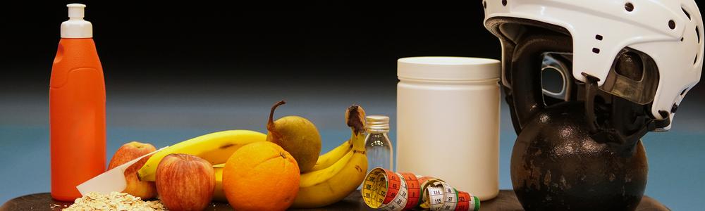 Arrangemang av tränings- och hälsorelaterade föremål på ett bord, inklusive vattenflaska, frukt, havregryn, kosttillskott, måttband samt kettlebell med hjälm.