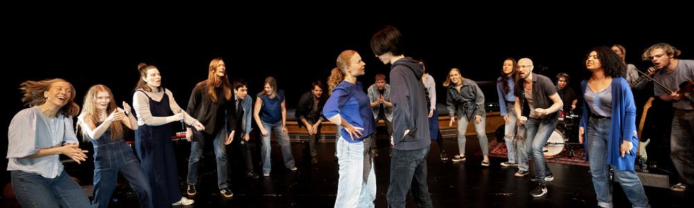 En grupp teaterlärarstudenter på scen i en halvcirkel. I mitten står två personer.