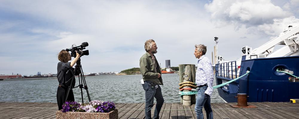 kvinna som lyssnar i hörlurar, man som fotograferar och en hög med tidningar