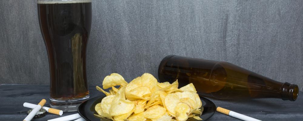 Unhealthy lifestyle concept, cigarettes, plate of crisps and beer on the dark background