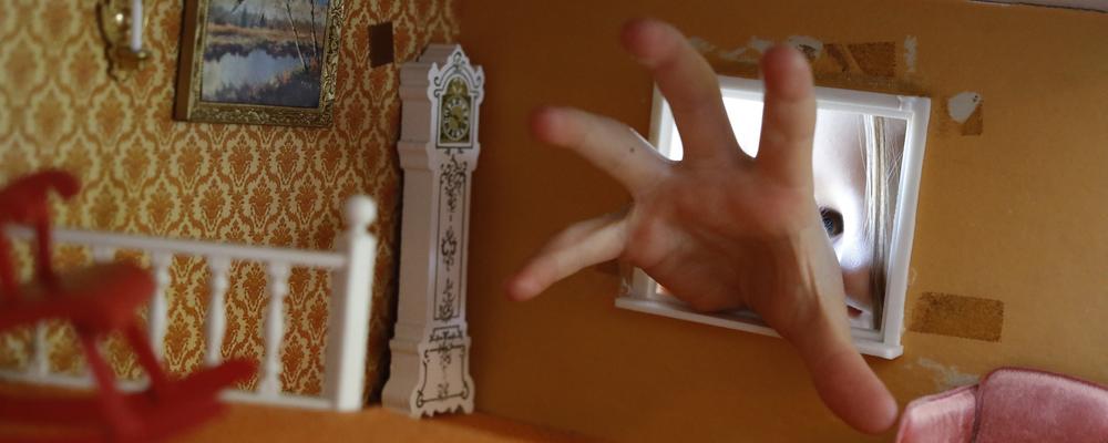 Bild på en hand som tränger sig in genom ett fönster i ett dockskåp