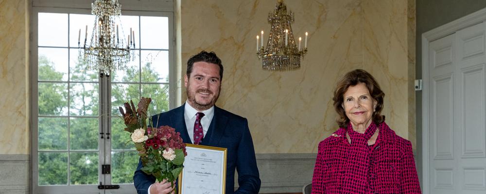 Nicholas Ashton med diplom och blommor brevid HM Drottning Silvia i slottssal