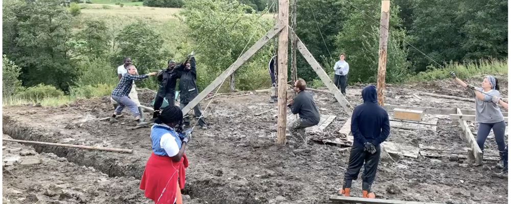 10 people pull ropes to jointly get a pole in place in the muddy ground. The trees are green and it is blowing in the treetops.