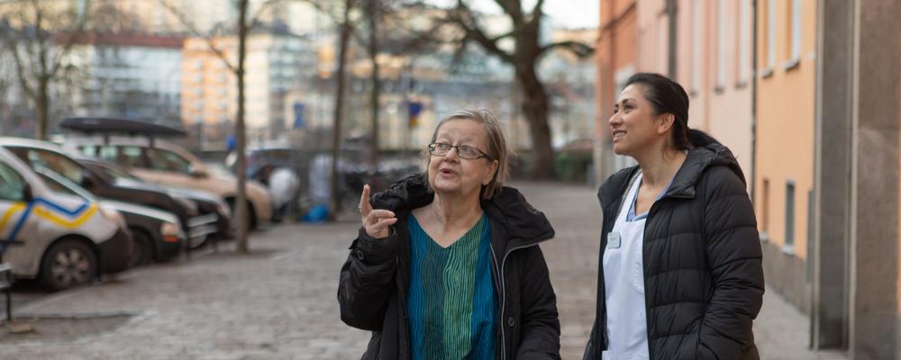En sköterska och en äldre kvinna står tillsammans på en gata och pratar.
