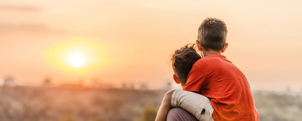 Ett barn sitter på sin pappas axlar, tillsammans tittar de mot solnedgången.