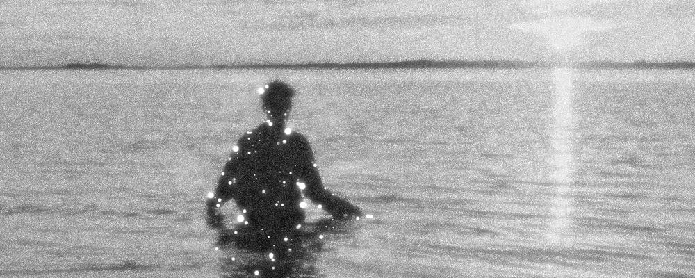 Person fotograferad i svartvitt motljus, med vatten till midjan. Havet glittrar.