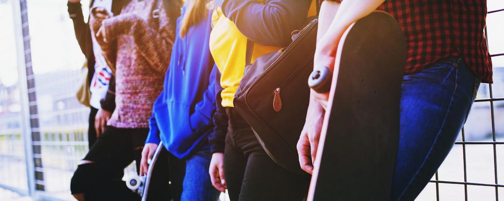 En grupp unga människor står tillsammans mot ett staket. En av ungdomarna håller en skateboard under armen.