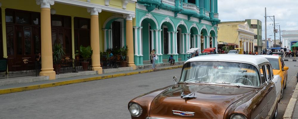 Äldre bil, glansigt brun, parkerad på en gata i Kuba med färgglada hus i grönt och gult. 