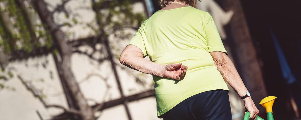 Äldre kvinna med ryggvärk vattnar växter i sin trädgård.