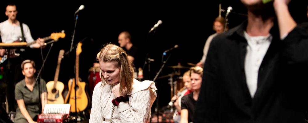 Students on stage during a performance. In the middle is a musical theatre student holding a phone.