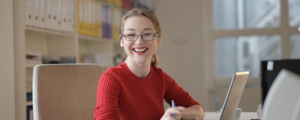 Kvinna som ler har en röd tröja och sitter vid ett skrivbord.