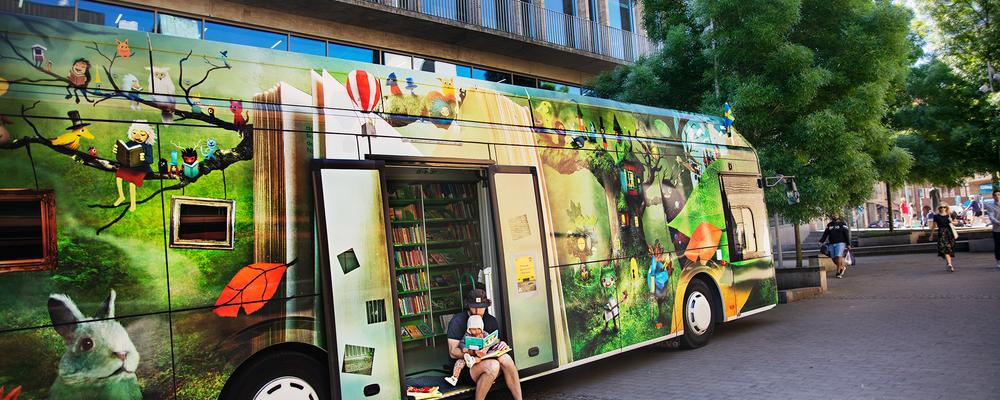 En av Göteborgs Stads eldrivna bokbussar står framför stadsbiblioteket i Göteborg. En pappa sitter i dörröppningen i och läser högt för sitt barn. En bokbuss är en buss som inretts till ett bibliotek och som kan köra runt till olika platser. 