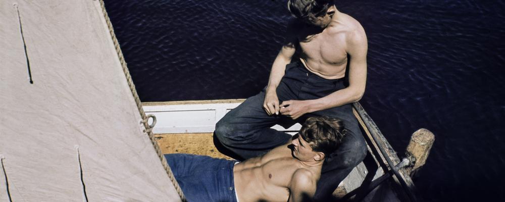 Two men are sitting in a boat. One is carefully cradled in the lap of the other.