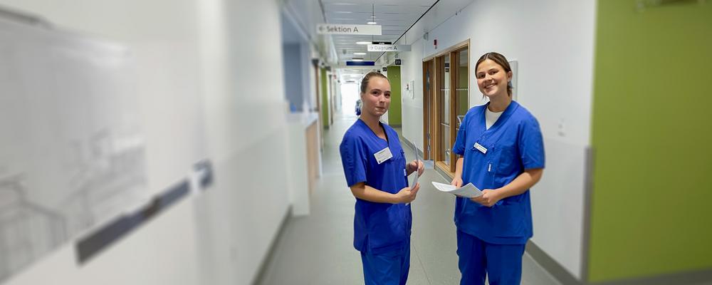 två kvinnliga studenter klädda i båa vårdkläder i en sjuhuskorridor