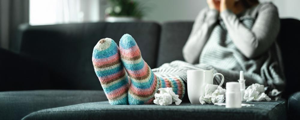 woman sitting on the couch blowing her nose, with tea and nasal spray, and antipyretic