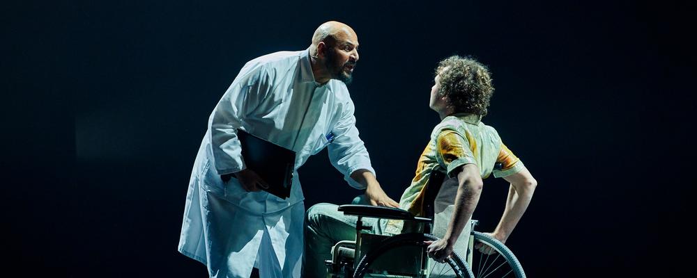 På secen står en läkare i vit rock lutad över en person i rullstol