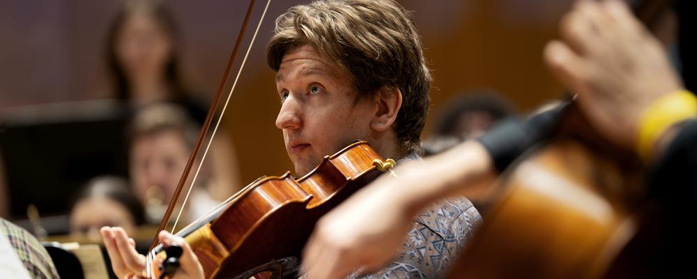 En person spelar fiol, andra musiker i oskärpa syns också