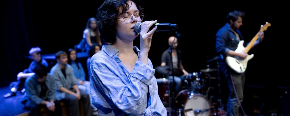 En person sjunger, i bakgrunden fler personer med musikinstrument