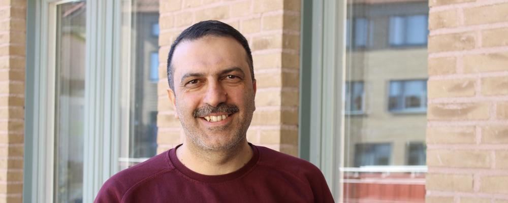 En man ler mot kameran och står på en uteplats med två fönster och tegelvägg bakom sig.