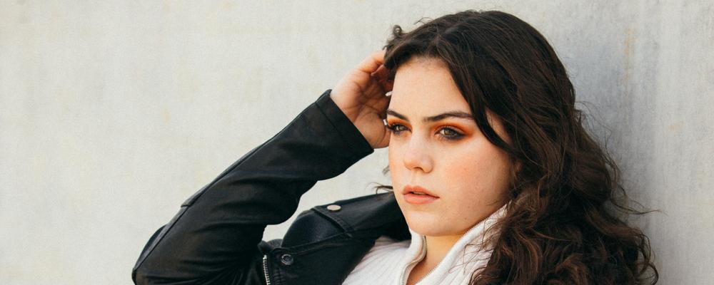 En tjej i tonåren står nära en vit husvägg och fixar lite med sitt hår med ena handen