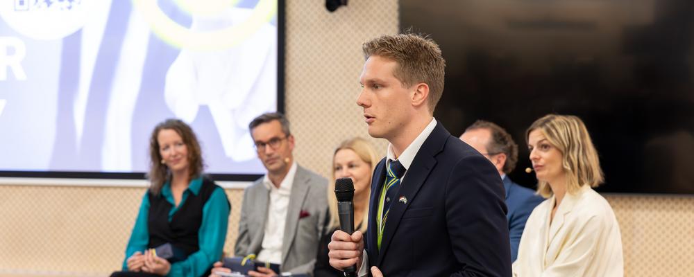 En person står och pratar, några sitter bredvid och lyssnar