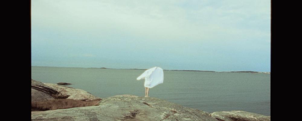 En person står på en klipphäll framför havet och svepet en vit handduk över axlarna