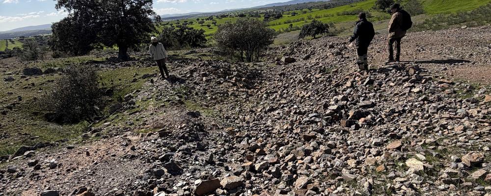 Tre personer står och tittar på en grushög i ett landområde med träd och blå himmel.
