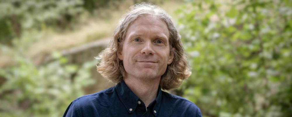 Porträttfoto på Mattias Svensson tagen utomhus med grönt lövverk i bakgrunden