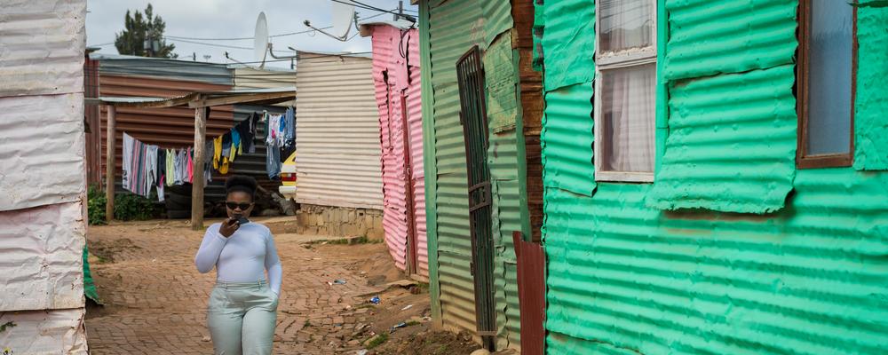 A person walking along a narrow street lined with colorful corrugated metal houses, with laundry hanging on a line in the background.