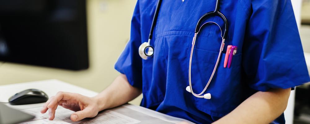 Healthcare worker in blue scrubs using laptop with stethoscope around neck