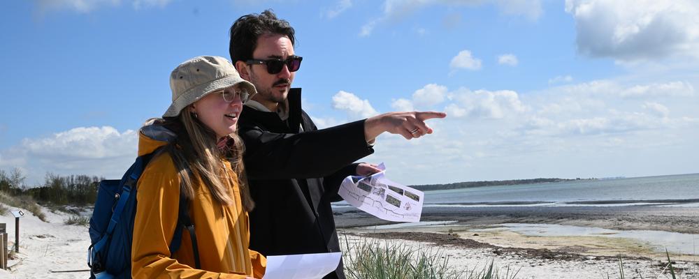 Två studenter står på en strand nära ett hav och gör fältstudier. 