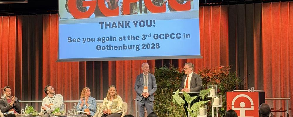 People sitting in chairs and two people standing on the conference stage. A screen is showing the words Thank you. 