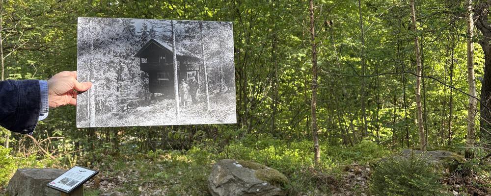 Kvarlämningar av en husgrund, i ett somrigt landskap. Någon håller in ett svartvitt fotografi över byggnaden som stod här tidigare. 