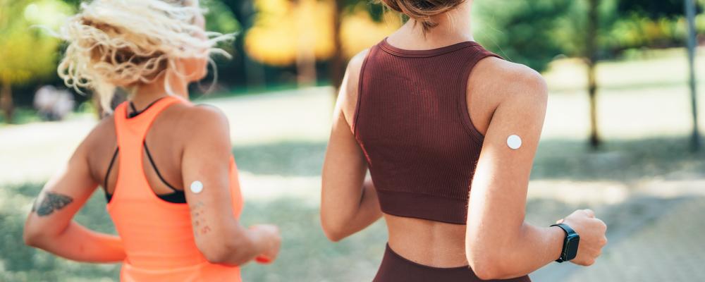 Two people jog in a park wearing sensors on their upper arms.