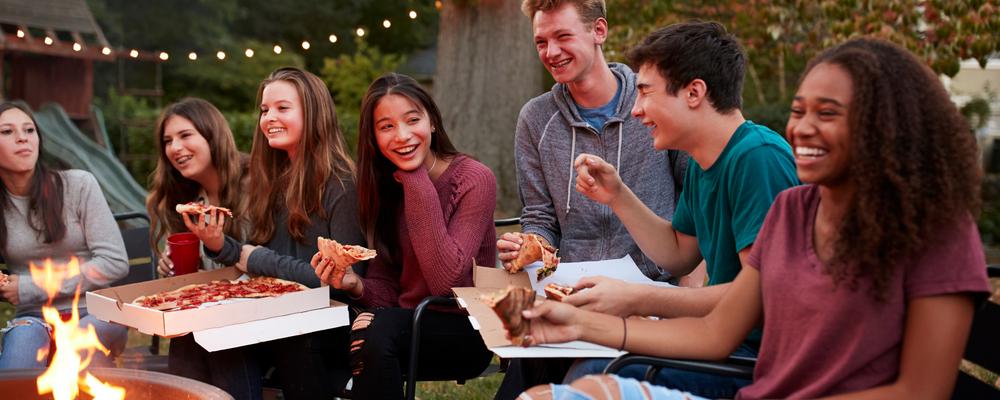 Bilden visar några ungdomar som sitter runt en eld och äter pizza och har trevligt tillsammans.