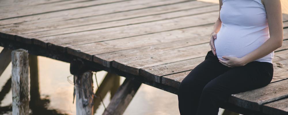 Gravid person sitter på en träbrygga med händerna på magen.