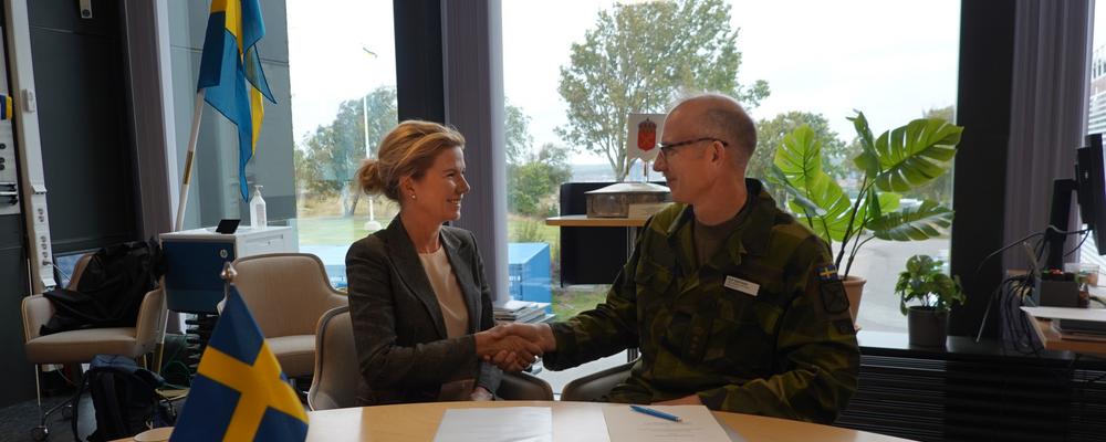 Two people shake hands at a table with Swedish flags.