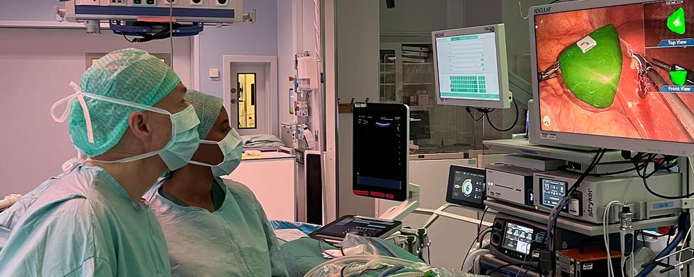  Två personer i operationskläder följer en skärm under en titthålsoperation. På skärmen syns en grön 3D-visualisering ovanpå kamerabilden av organet.