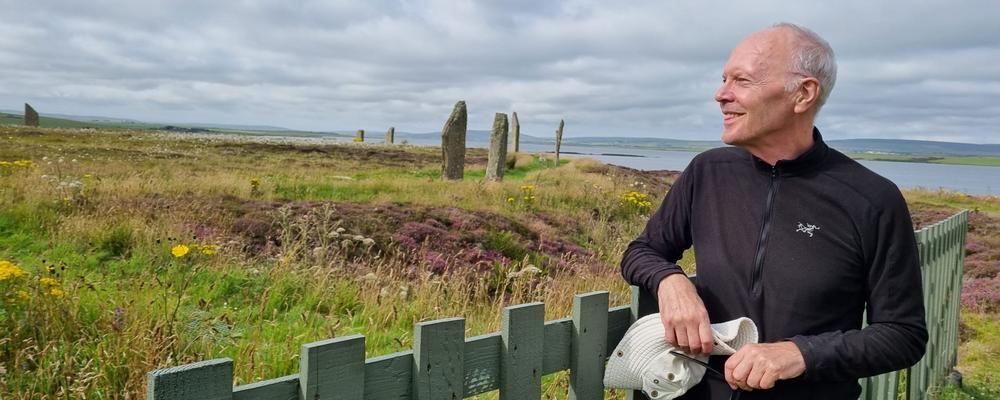 A man with grey hair in black leisurewear is standing to the right, leaning to a green wooden fence and looking out over a field. He smiles. On the field, there are some vertical stones, the Ring of Brodgar. Behind him a lake area.