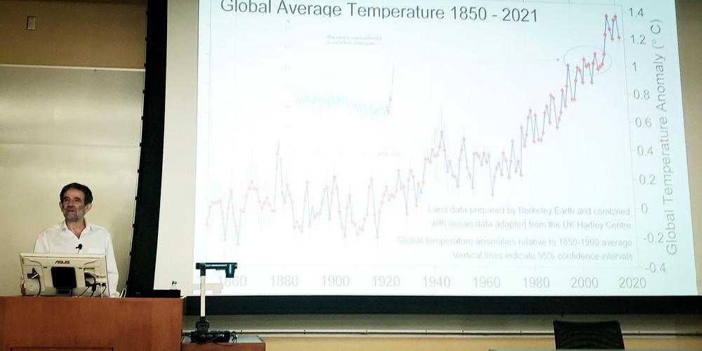 En man med mörkt hår föreläser framför en projektion av en graf över jordens medeltemperatur sedan 1850.