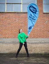 Alice i green sweater holding a flag with the HUJ logo