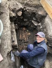 En kvinna sitter i en grävd grop med en arkeologisk spade i handen.